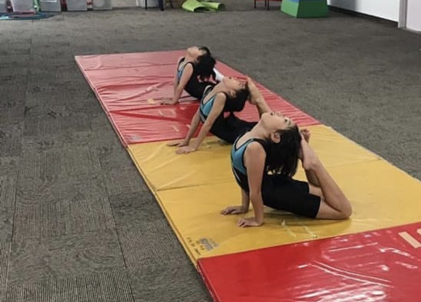 Three gymnasts practising floor stretches on red and yellow mats.