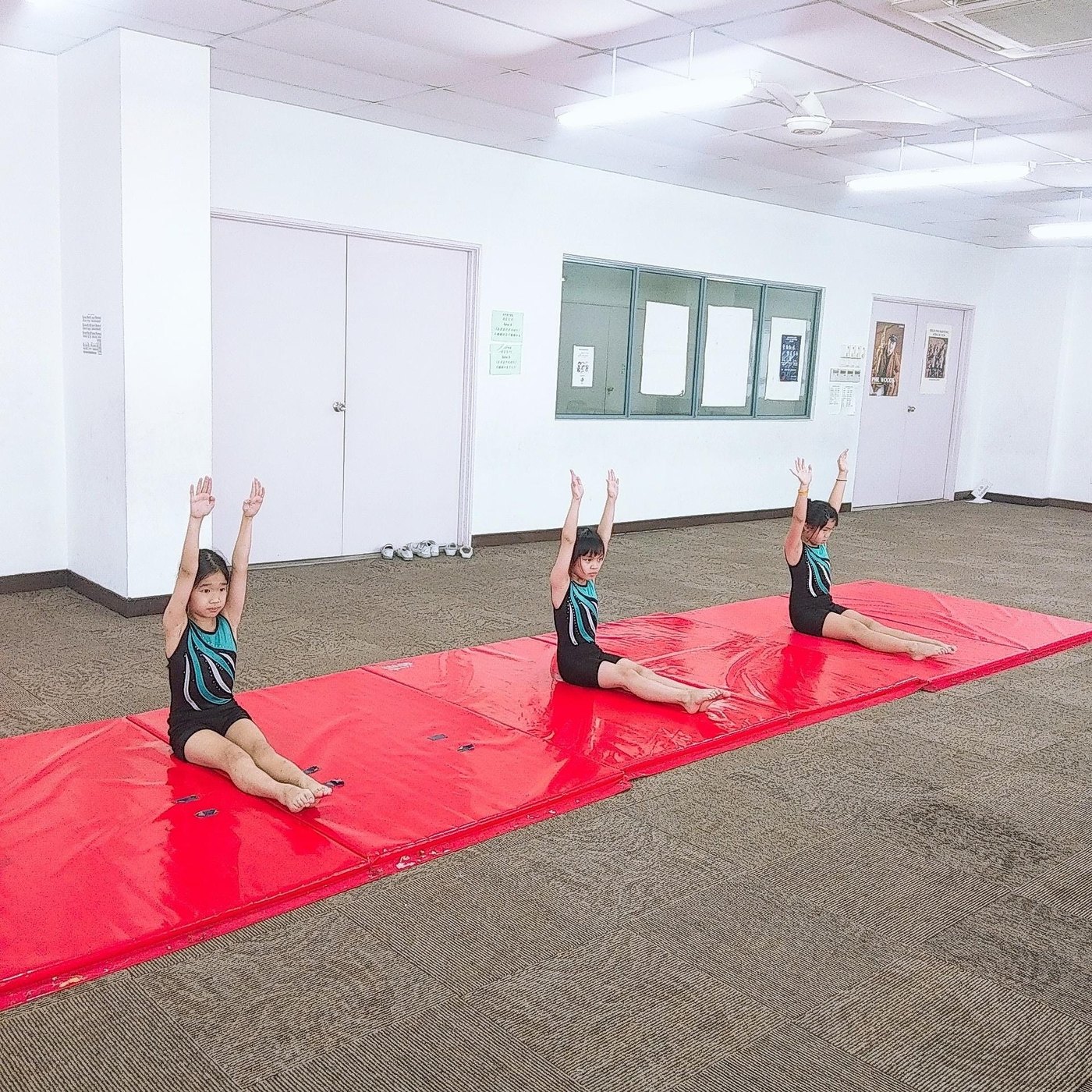 Three students practising seated posture and arm positions on red gymnastics mats.